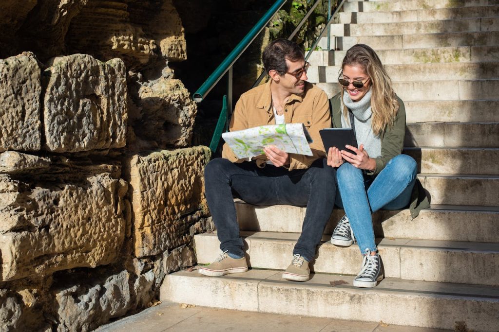 Smiling couple explores city sitting on stairs with map and tablet.